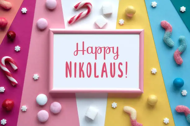 Text Happy Nikolaus in pink frame. Large heart lolly pop, assorted sweets, chocolates, confectionery on multicolor layered paper. Flat lay, top view. Greeting design for December traditional holiday.