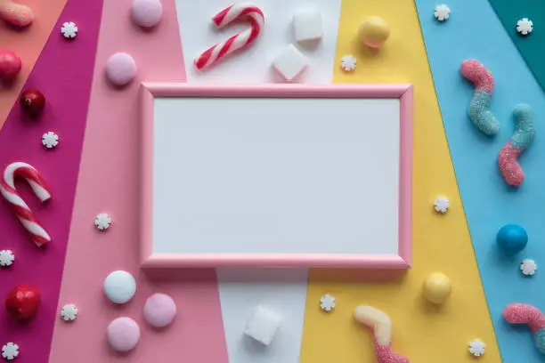 Blank pink white frame on layered colored paper. Assorted sweets, confectionery, chocolates and candy canes around empty frame. Copy-space, place for text. Flat lay, top view. Colorful greeting design.