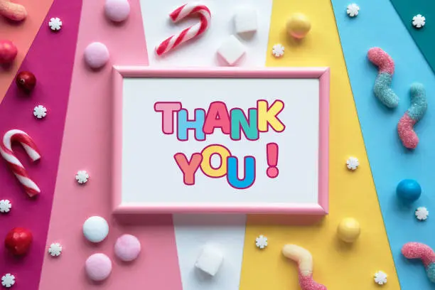 Text Thank you in frame on layered colored paper. Assorted sweets, confectionery, chocolates and candy canes around the frame. Flat lay, top view. Colorful greeting design.