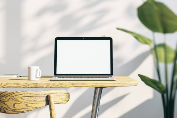 istock Close up of creative designer office desktop with empty white laptop screen with mock up place in frame, concrete wall background with sunlight and shadows, blurry decorative plant and coffee cup. 3D Rendering. 1435106124