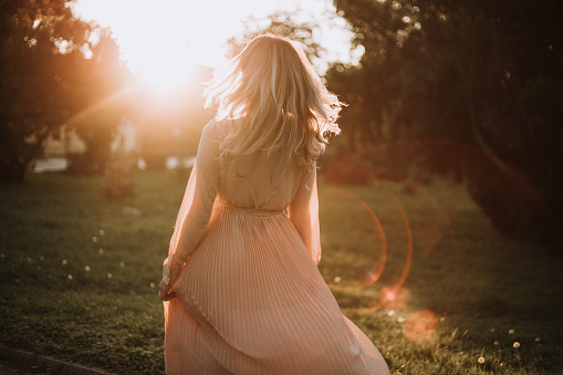 Beautiful young woman in pink dress standing in front of city park. Long hair with little makeup and few rays of sunshine. Nature and autumn idyll.