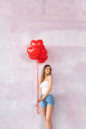 Valentine smiling girl holding red air balloons, posing in pajamas on the pink wall. Beautiful Happy Young woman. A lot of copy space.