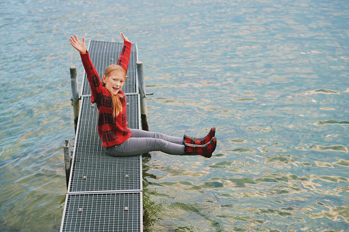 Outdoor portrait of cute little girl posing next to lake on dock, wearing red sweater and rain boots, cold fall or spring weather