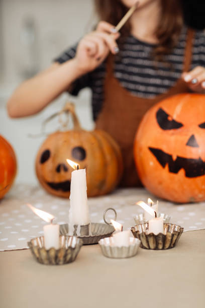 une petite fille allume une bougie pour halloween. petite fille en costume de sorcière avec citrouille sculptée avec un visage fait par un enfant. joyeuse famille se préparant pour halloween. mise au point sélective - jack fruit photos et images de collection