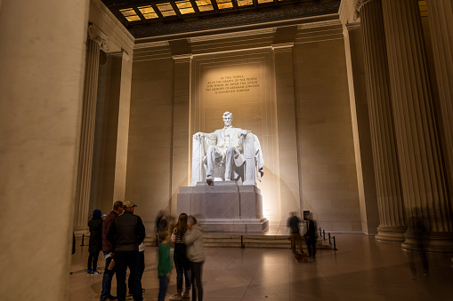 Tourists visit the Lincoln Memorial in Washington, D.C. at night on a winter night. A long exposure shows blurred people beneath the impressive statue of President Abraham Lincoln, sitting in a chair.