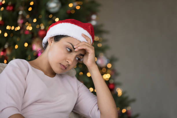Depressed frustrated Indian girl in Christmas hat going through problems Depressed frustrated Indian girl in Christmas hat going through problems, emotional crisis, negative mood, feeling apathy, loneliness, sitting on sofa at Xmas tree lights, thinking over bad news asian christmas stress stock pictures, royalty-free photos & images