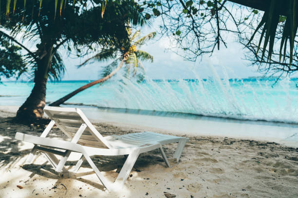 A single white beach daybed in front of a waterfront with splashes of turquoise water; an empty recliner on the coastline of a luxury tropical resort with a splatter of teal waves crashing on the pier