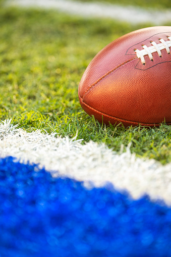 A low angle view of a leather American Football spotted next to a goal line within inches of a blue end zone.