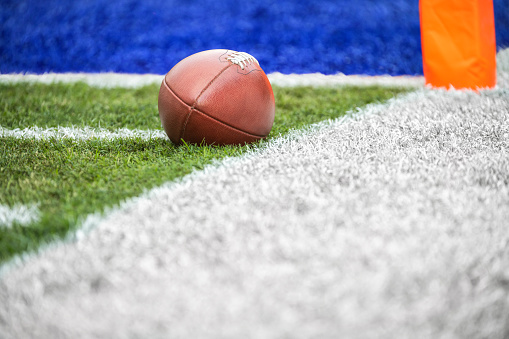 A low angle view of a leather American Football spotted next to a white side line within a yard of a blue end zone.