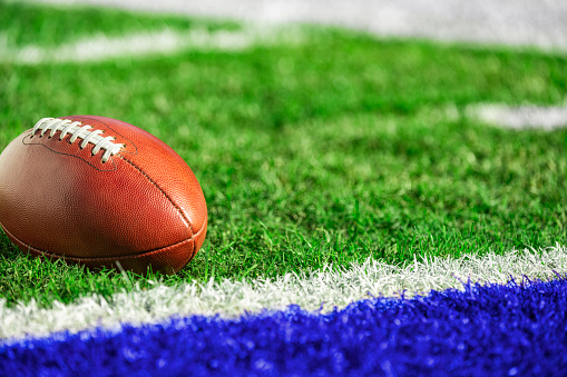 A low angle view of a leather American Football spotted next to a goal line within inches of a blue end zone.