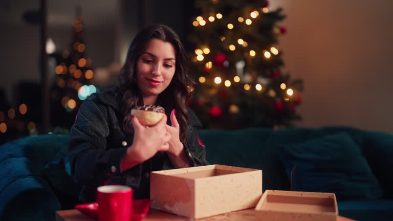 Young woman opening gift/subscription box and enjoying eating doughnut while relaxing in living room at home during Christmas