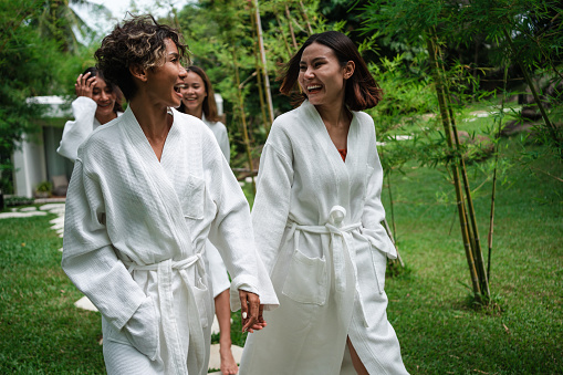 Smiling women are walking in bathrobes to the sauna through the green yard.