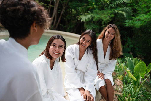 Smiling women are sitting in bathrobes by the small pool after the sauna on their vacation in a luxury villa.