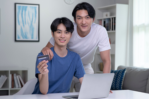 Young Asian gay couple smiling looking at camera while holding credit card and using laptop computer. LGBT gay businessman working at home. Online shopping, e-commerce, internet banking, spending money