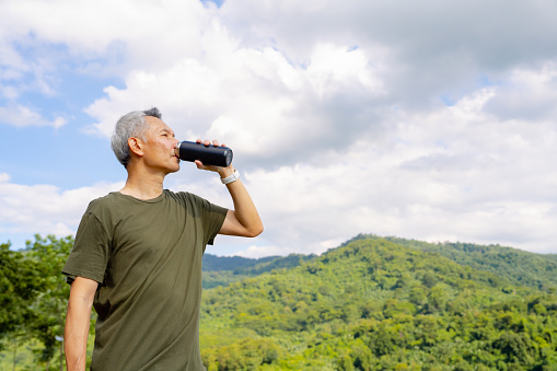 Active mature man drinking water from reusable packaging while at relaxing on bluff the mountains after exercise.