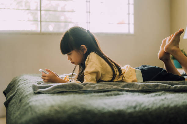 une écolière de 8 ans est allongée sur son lit en regardant le téléphone, en envoyant un message texte dans une application mobile sur un smartphone après l’école - female 8 9 years child excitement photos et images de collection