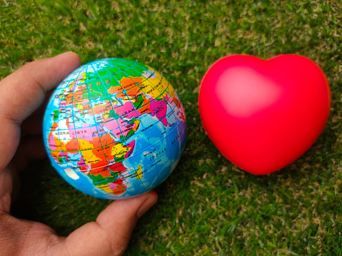 Hand hoding the earth globe with red heart on a green grass background.