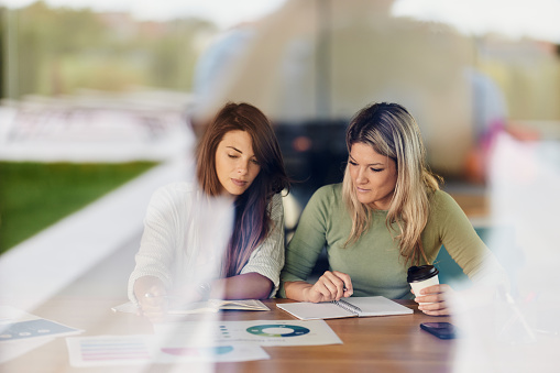 Young creative women cooperating while examining business charts in the office. The view is through glass.