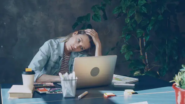 Young blond woman is sitting in office working with computer. She is tired after long day at work.