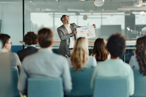 Mature male leader talking to large group of his colleagues about business reports on a seminar in board room.