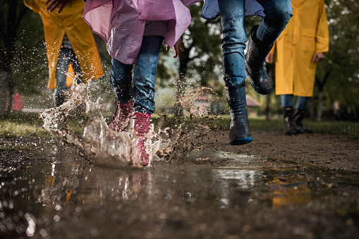 Unrecognizable siblings having fun while running through muddy puddle in the park. There are people in the background.