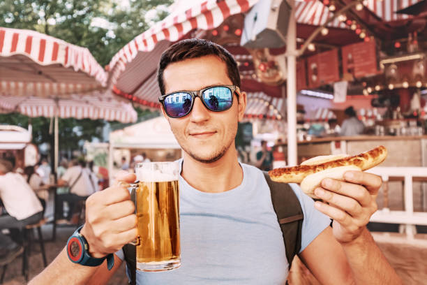 Happy man drinking beer and eating traditional german bratwurst - hotdog at funfair and street food festival. National cuisine and biergarten concept stock photo