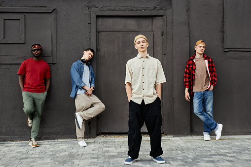 Diverse group of boys wearing street style clothes standing against black wall outdoors and looking at camera, full length portrait