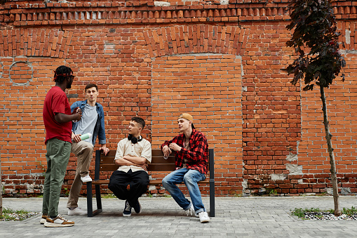 Wide angle view at diverse group of young guys wearing street style fashion in urban city area against brick wall and chatting, copy space