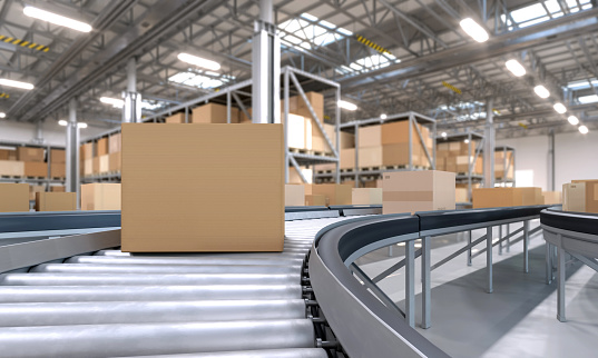 Close-up of blank cardboard box moving on a conveyor belt in a large distribution warehouse with shelves full of boxes, brightly illuminated. Conveyor belt point of view, with copy space on package side for adding custom graphics.
