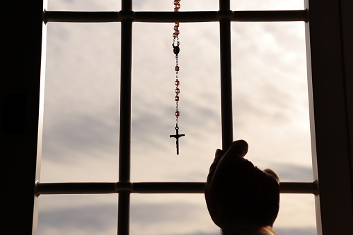 Person in the act of prayer in front of the rosary hanging from a window with a grate.