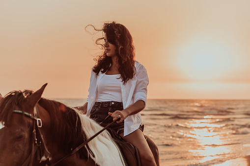 A woman in summer clothes enjoys riding a horse on a beautiful sandy beach at sunset. Selective focus. High quality photo