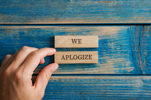 Male hand assembling a We apologize sign written on two wooden pegs placed over blue wooden background.