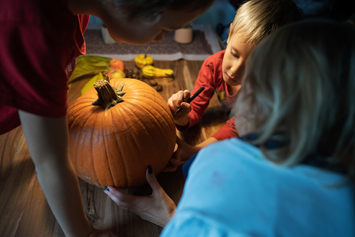 Three children, siblings, drawing on a pumpkin they will carve for halloween holiday season.