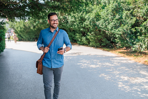 Smiling adult bearded man, carrying his phone and handbag, enjoying a beautiful day outside.