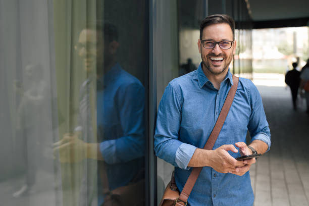 Portrait of delighted entrepreneur businessman, smiling for the camera, outdoors. Picture of happy adult man, posing for the photo, using his phone outside the building. businessman happiness outdoors cheerful stock pictures, royalty-free photos & images