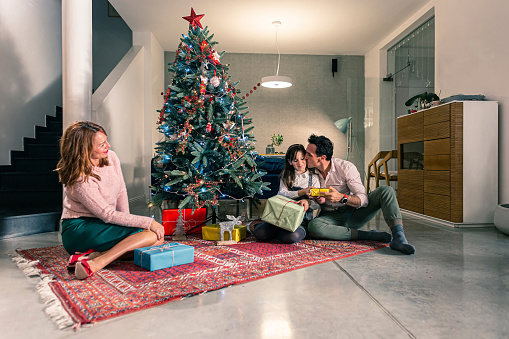 A mother and father sitting with their daughter on the floor next to the Christmas tree and exchanging Christmas presents. The living room is decorated in the spirit of the upcoming holidays.