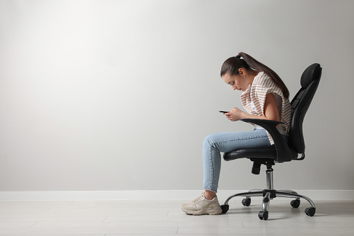 Young woman with poor posture using smartphone while sitting on chair near grey wall, space for text