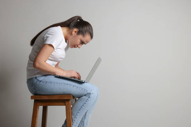 istock Young woman with poor posture using laptop while sitting on stool against grey background, space for text 1435292770
