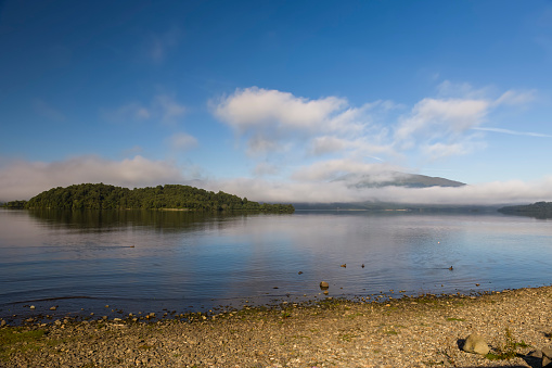 Early morning view of tree covered Inchlonaig Island over a calm Loch Lomond from the beach at Cashel in Scotland, UK