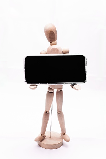 A vertical shot of a wooden doll on a base holding a smartphone with black screen on a white background
