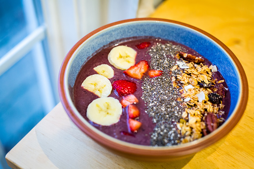 Healthy and nutritious Acai Bowl made with Acai, coconut milk, coconut yogurt, with additional toppings of chia seeds, granola, sliced bananas, strawberries, pecans, dried fruit and coconut flakes  on a table