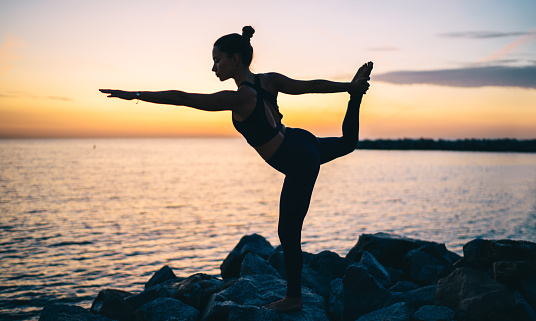 Sporty female doing yoga Natarajasana asana while practicing balance during training on beach in evening in nature while enjoying time