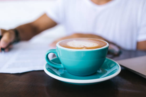 istock Selective focus on blue cup with caffeine beverage standing on working place with milky foram with coffee art on top, cropped image of hot coffee standing on blurred background with writing hand 1435316483