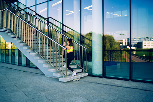 Young female in sports clothing and white sneakers doing warm up lunge exercise on outside stairs of building on sunny morning