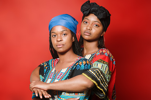 Two black afro latina women with a serious expression on their face looking at the camera. They are wearing colorful clothes and are standing in front of a red background.
