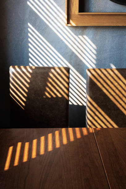 Light and shadow from the window. Thin lines of light on the wall, armchairs and table. Shadow from the blinds. Brown wall and table in an empty room