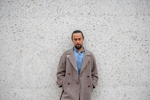 Portrait of hispanic adult male leaning back on large grey wall. He has dark hair and well-groomed beard. He is wearing a coat and blue shirt underneath.