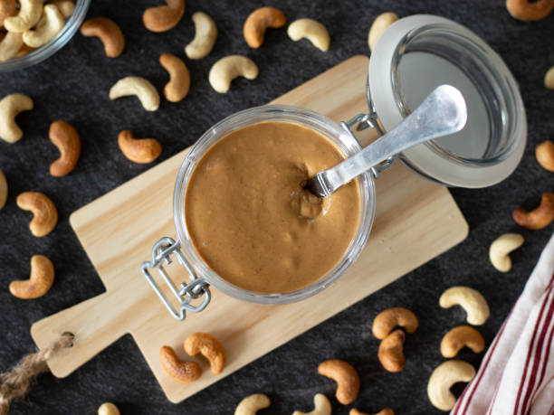 Homemade cashew butter in a jar with a knife and roasted and raw cashew nuts on dark background stock photo