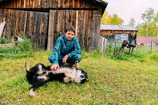 Copy Space Wide Angle Portrait of a Cute Stoic Male Caucasian Teenage Rancher Wearing a Button Down Shirt Embracing His Family Dog in Front of a Large Wooden Work Shed a Small Town Family-Owned Ranch in Colorado, USA