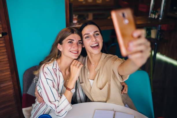 istock Caucasian influence bloggers looking at smartphone camera and smiling while clicking selfie images, happy female millennials 20s making cellphone images during together leisure in coffee shop 1435336285